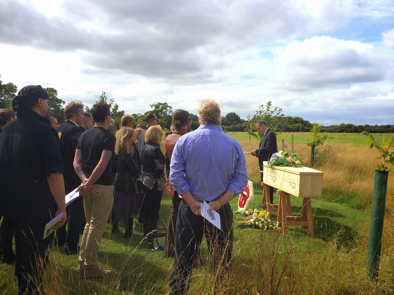 Humanist celebrant standing next to a pine wood coffin in the middle of a meadow with a group of mourners facing him while talking