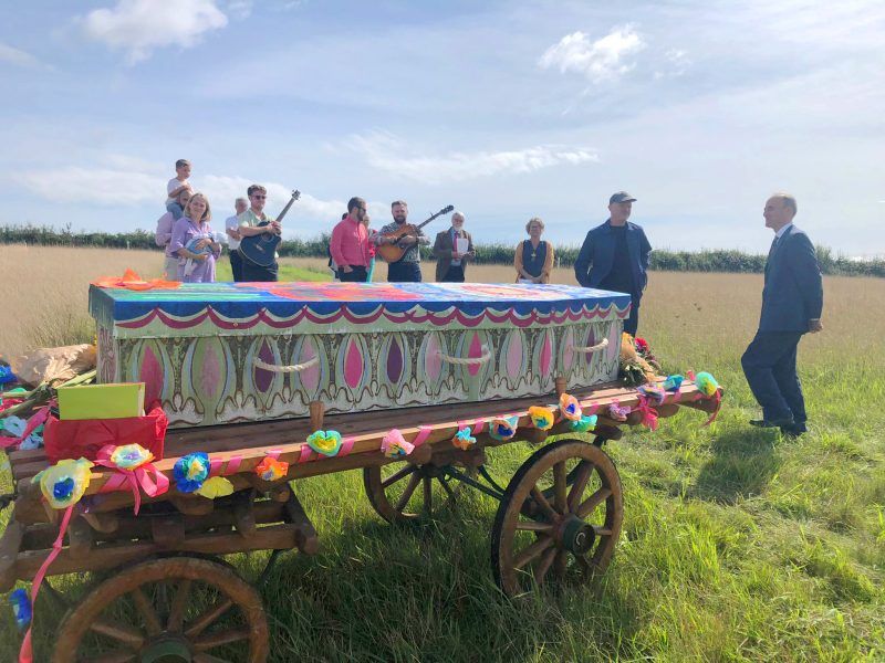 Celebration of life at Dorst Downs natural burial ground with a colourfully painted cardboard coffin and an attendee playing the guitar in the background