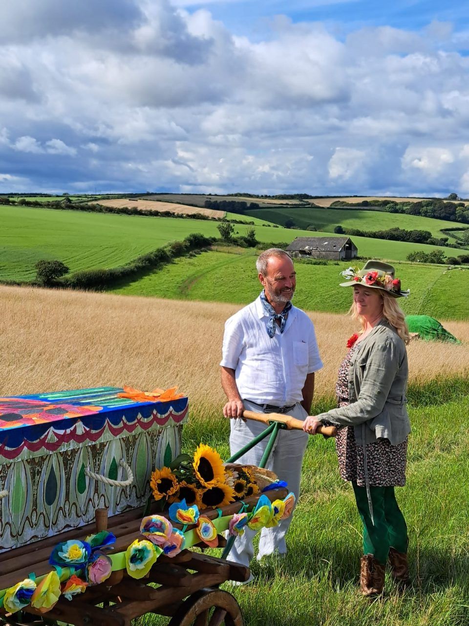 Celebration of life at Dorst Downs natural burial ground with a colourfully painted cardboard coffin on the wooden bier, with the rolling hills of Dorset downland behind.